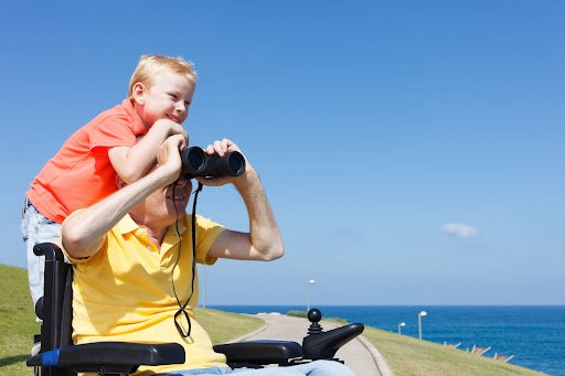 Disabled father and able child use binoculars' at the coast.